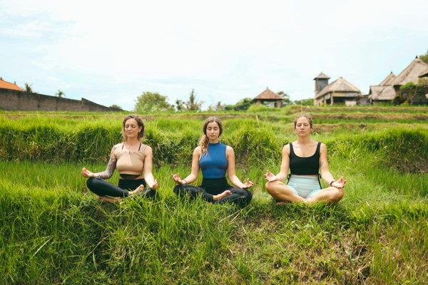 yoga small groups - Shades of Yoga Three women in athletic wear sit cross-legged, meditating on a grassy terraced hillside in a rural village setting.