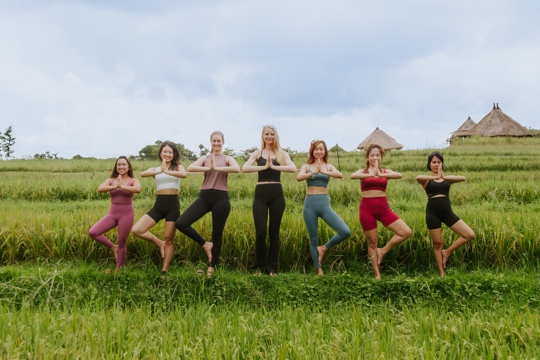 Eight women in colorful athletic wear perform yoga poses together in a grassy field with thatched huts in the background. - OR - Eight women practicing yoga outdoors in a green field, hands in prayer pose, huts visible in distance.